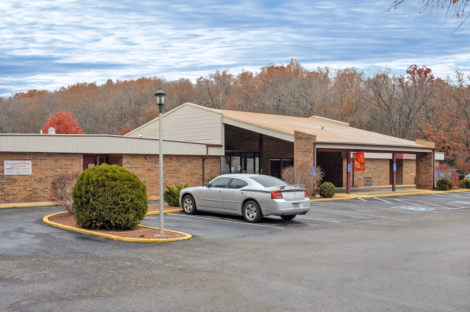 Exterior view of a single-story brick building with a sloped roof, surrounded by a parking lot with a silver car parked near the entrance. There are some bushes and trees with autumn foliage in the background under a partly cloudy sky.