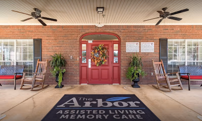 Red front door with wreath on brick porch, surrounded by rocking chairs and plants.