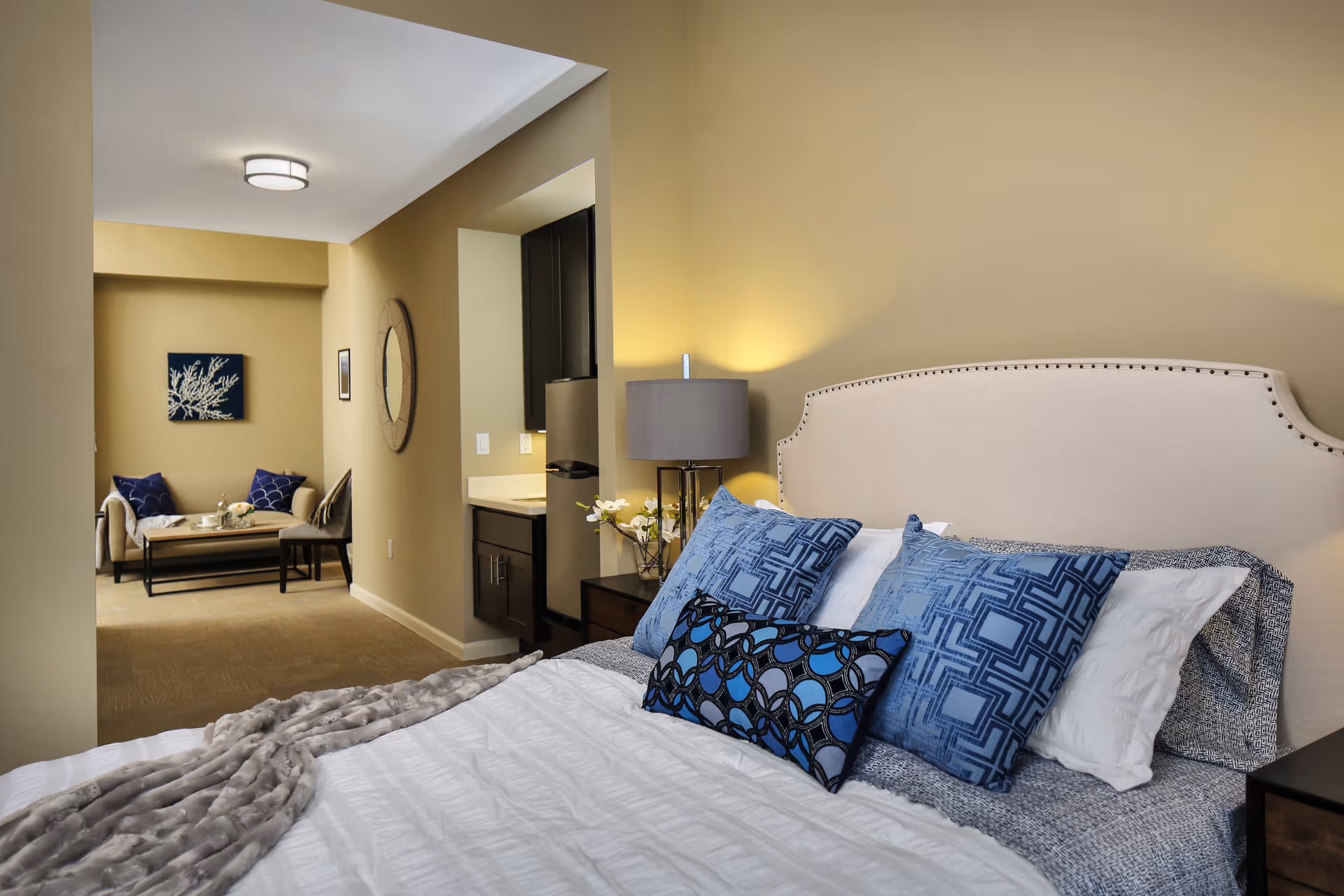 Modern bedroom with upholstered headboard, blue pillows, and a connected seating area and kitchenette.