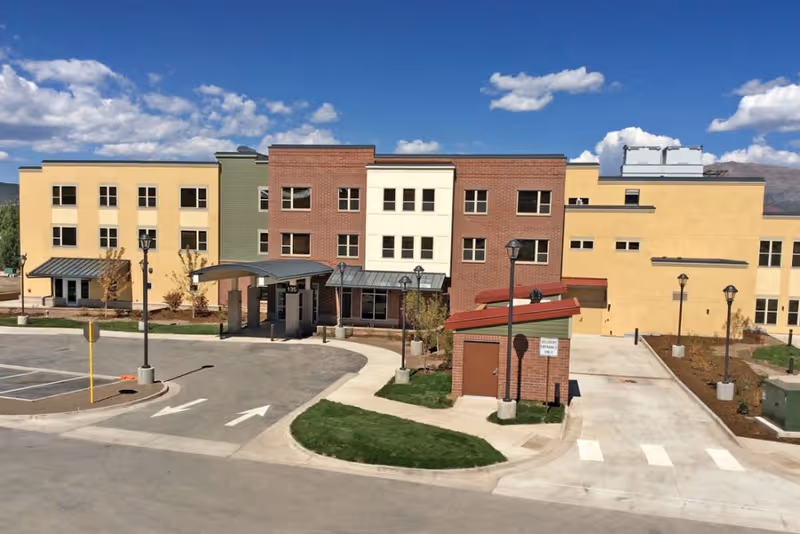 Exterior view of Castle Peak Assisted Living facility showing a multi-story building with yellow and brick facade under a blue sky with scattered clouds. The building has multiple windows, a covered entrance, street lamps, a parking area, and landscaped green patches.