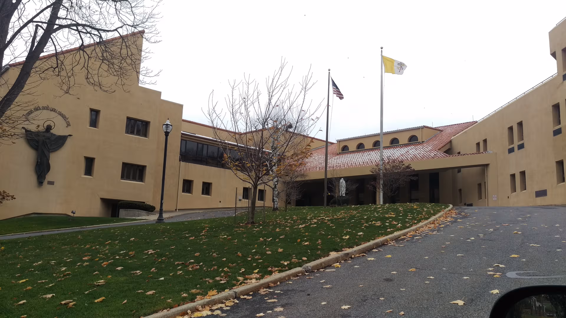 Exterior view of Rosary Hill Home showing beige building, flags, trees, and a curving driveway.