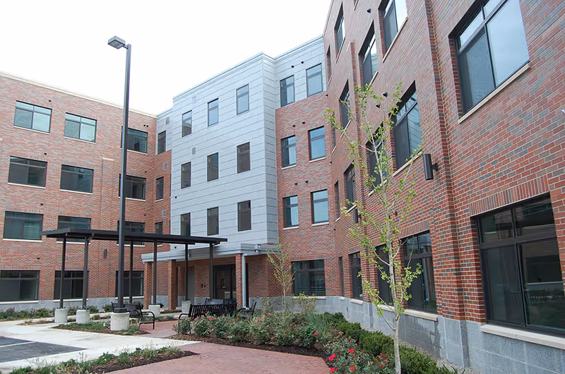 Courtyard and entrance of a multi-story brick assisted living building with benches, landscaping, and a covered walkway.