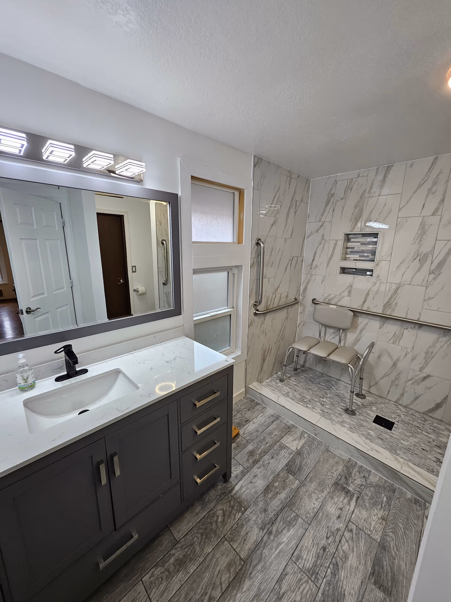 Modern bathroom with gray vanity, large mirror, accessible shower, and shower chair.
