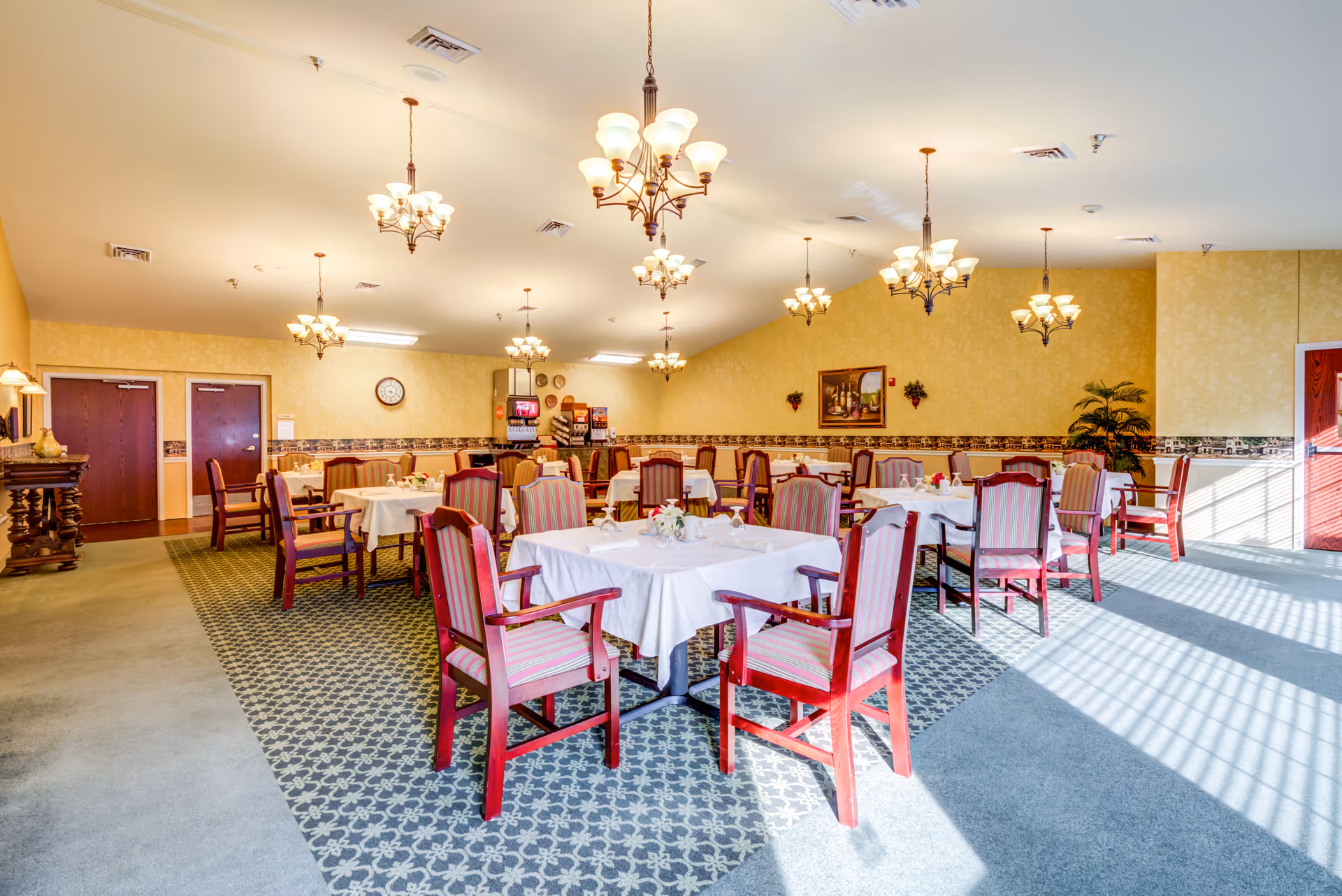 Bright dining room with round tables, white tablecloths, red chairs, and chandeliers.