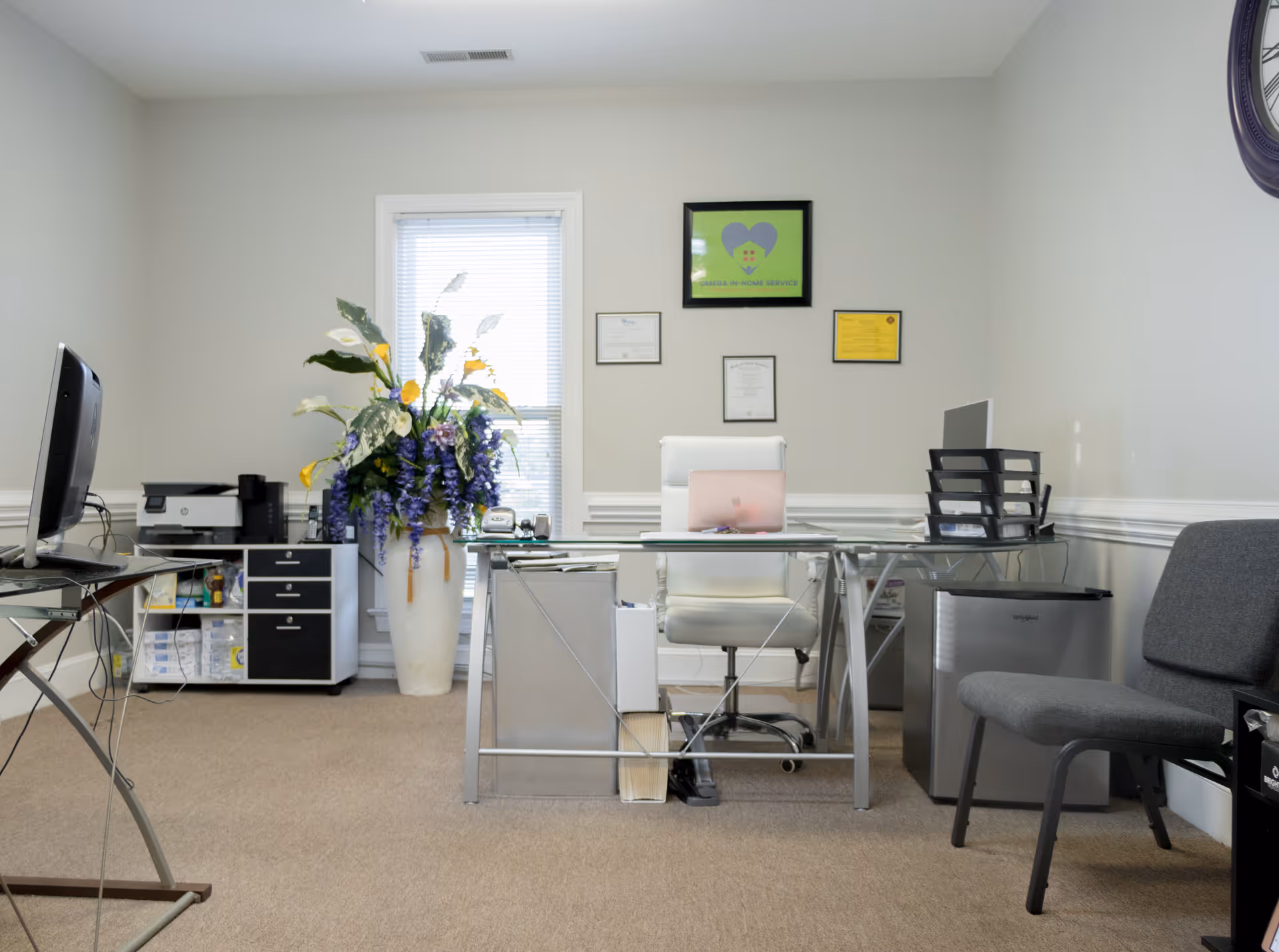 An office area in a senior living facility with a glass desk, laptop, plants, and certificates.