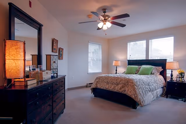 A cozy bedroom with a large bed featuring a patterned comforter and two green pillows. The room has two windows with blinds, two bedside tables each with a lamp, and a dark wooden dresser with a large mirror and a decorative lamp on top. The ceiling has a fan with lights.