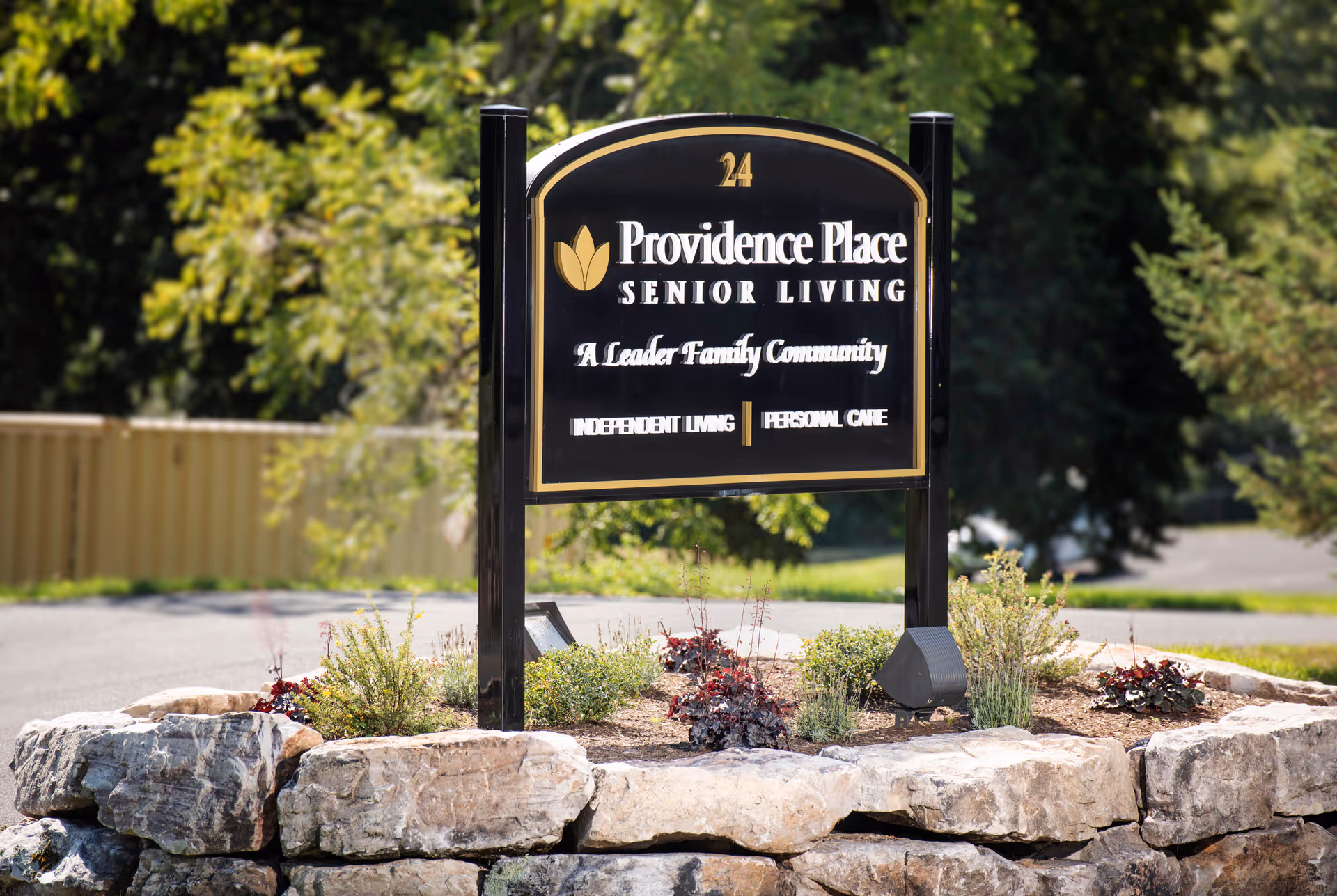 Black and gold entrance sign reading 'Providence Place Senior Living' mounted in a landscaped stone bed with trees in the background.