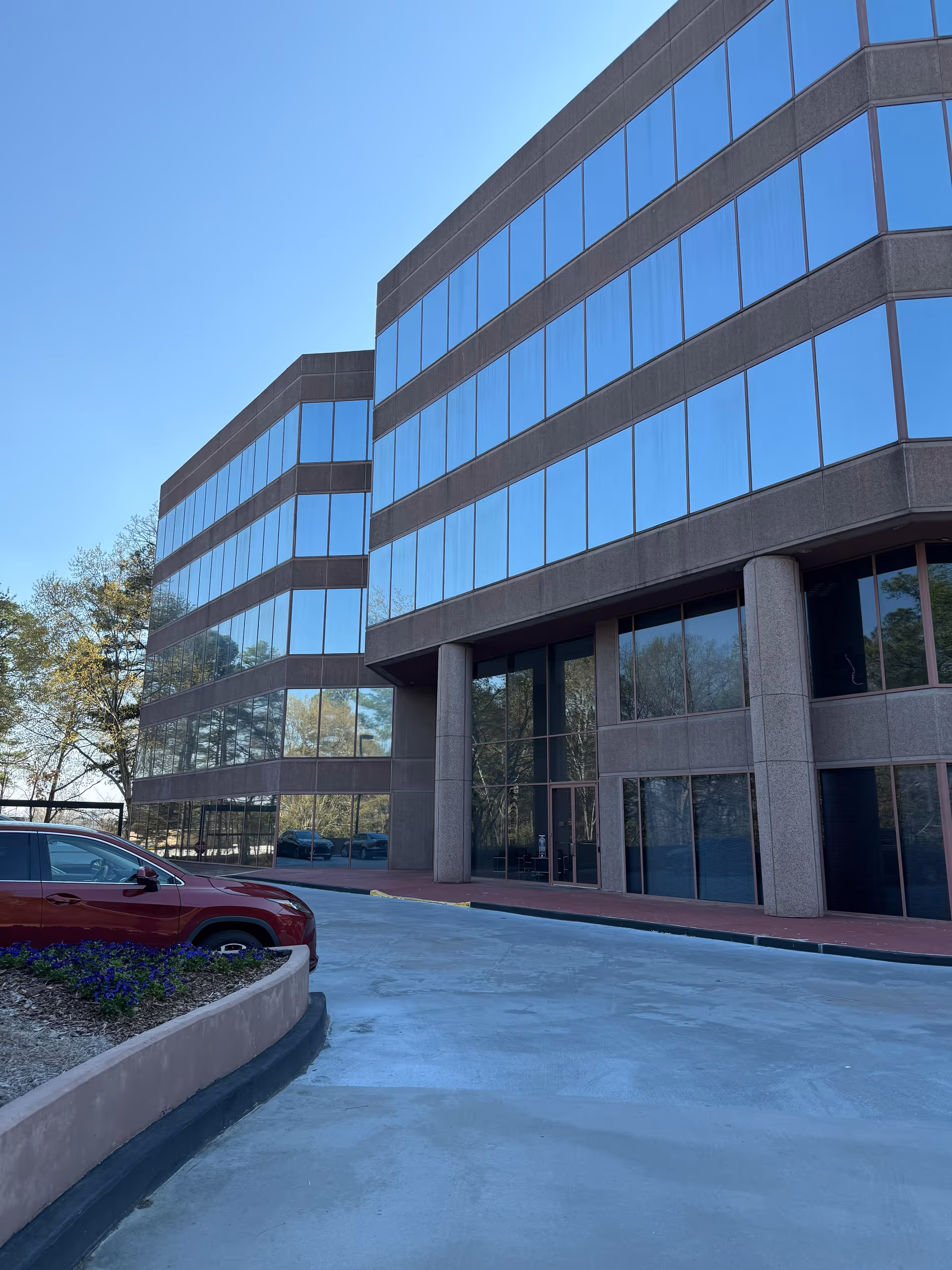 Modern glass front building exterior with curved facade, driveway, and a red car parked.