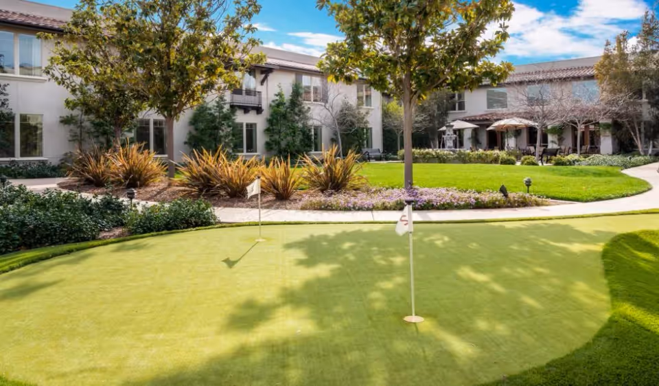 A landscaped courtyard featuring a small putting green with flags, trees, and a two-story building in the background.