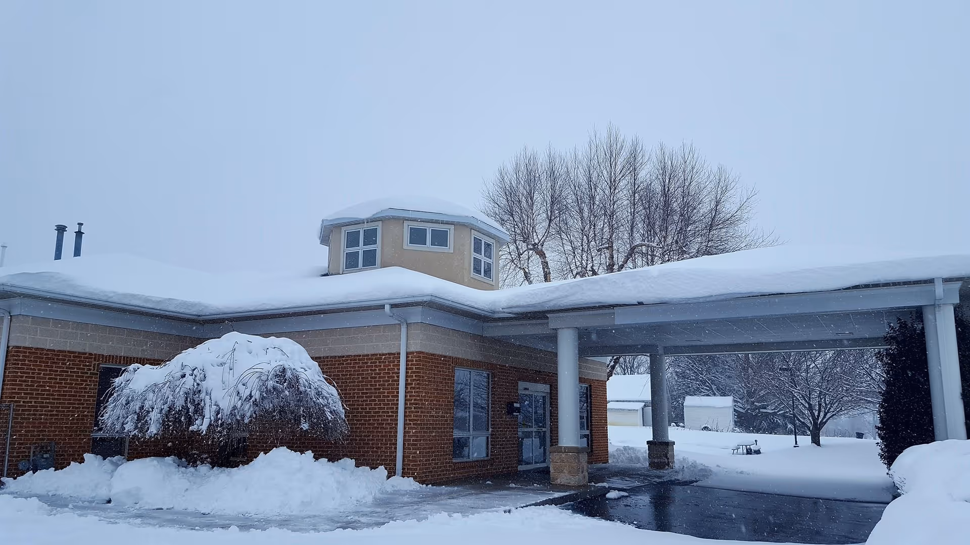 Snow covers the brick facility exterior and covered entrance with bare trees nearby.