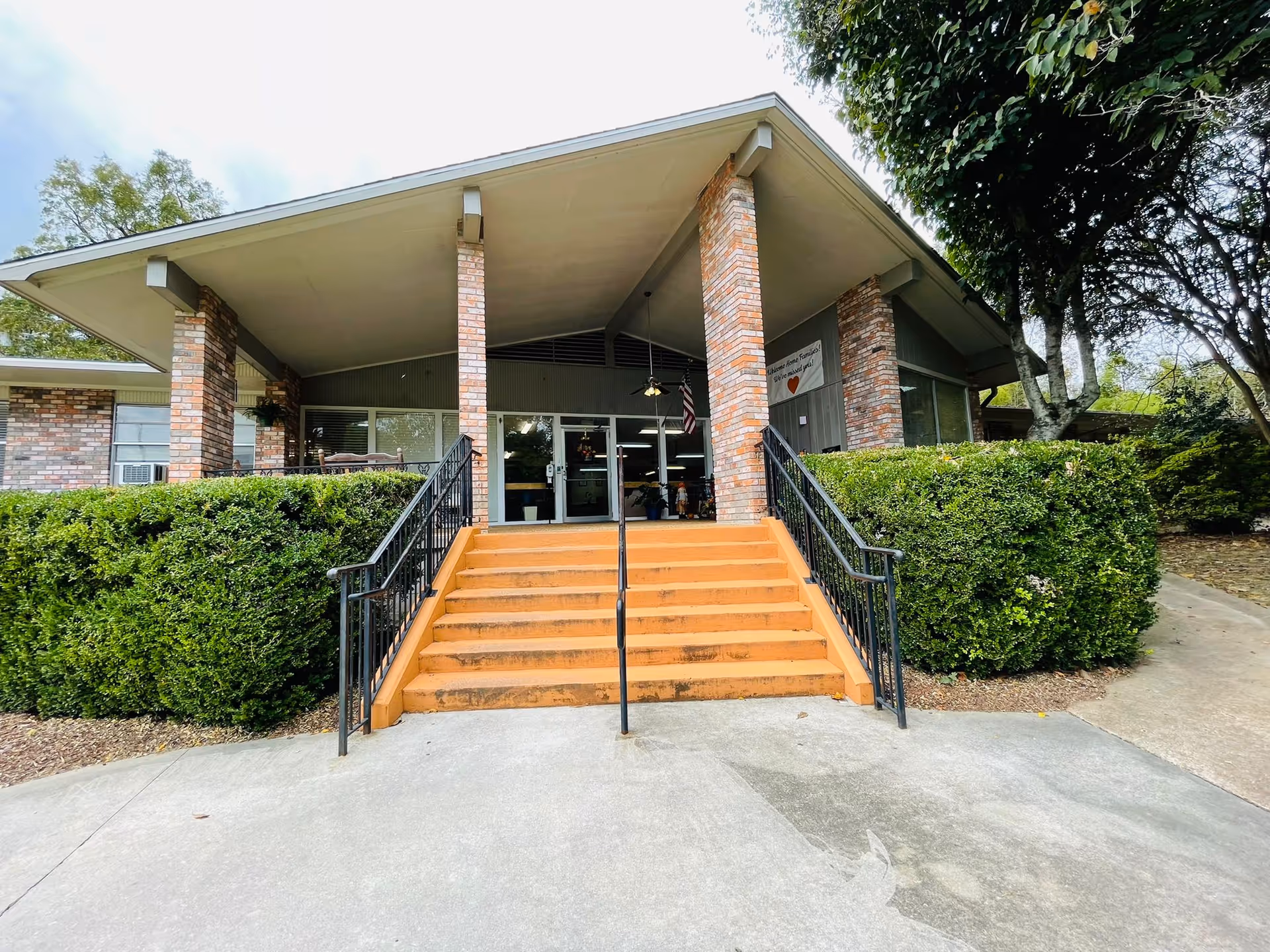 Front entrance of Natchez Rehabilitation and Healthcare Center showing a set of orange stairs with black railings leading up to double glass doors. The building has brick pillars and is surrounded by green bushes and trees.
