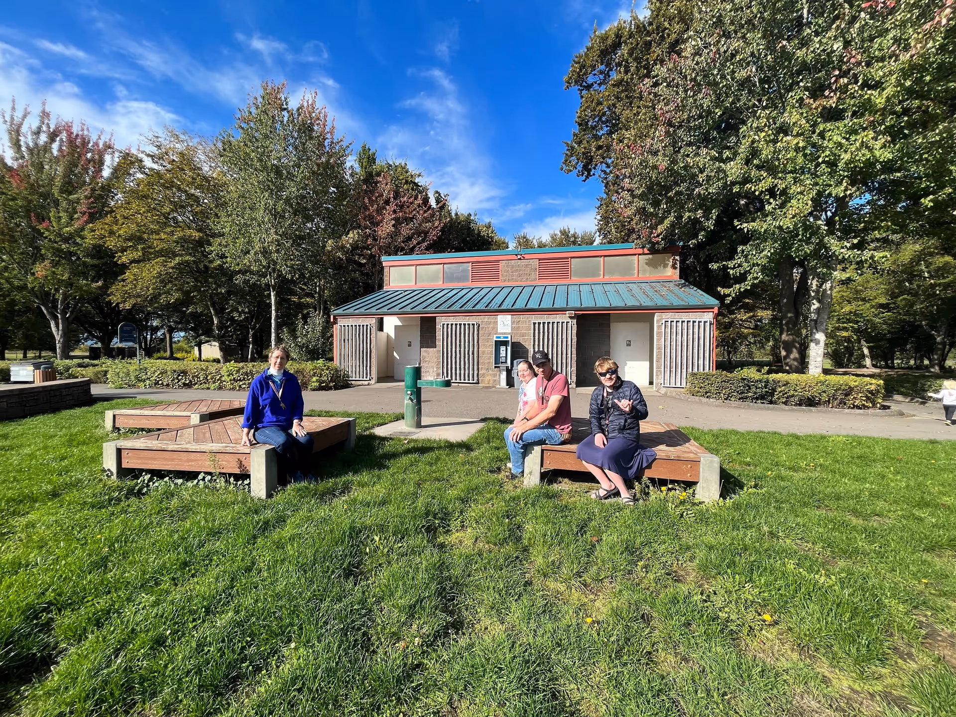 Group of people sitting on wooden benches in a grassy outdoor area with a building behind.