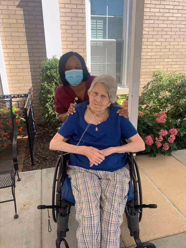 Caregiver wearing mask stands beside an elderly woman in a wheelchair on an outdoor patio.