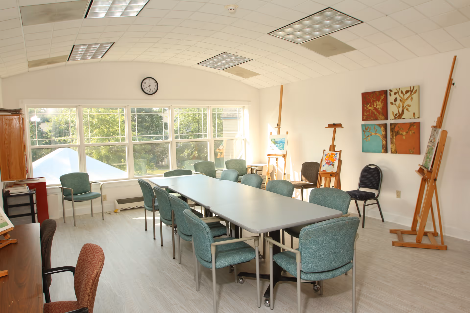 Bright activity room with a long table, teal chairs, easels, and wall art.