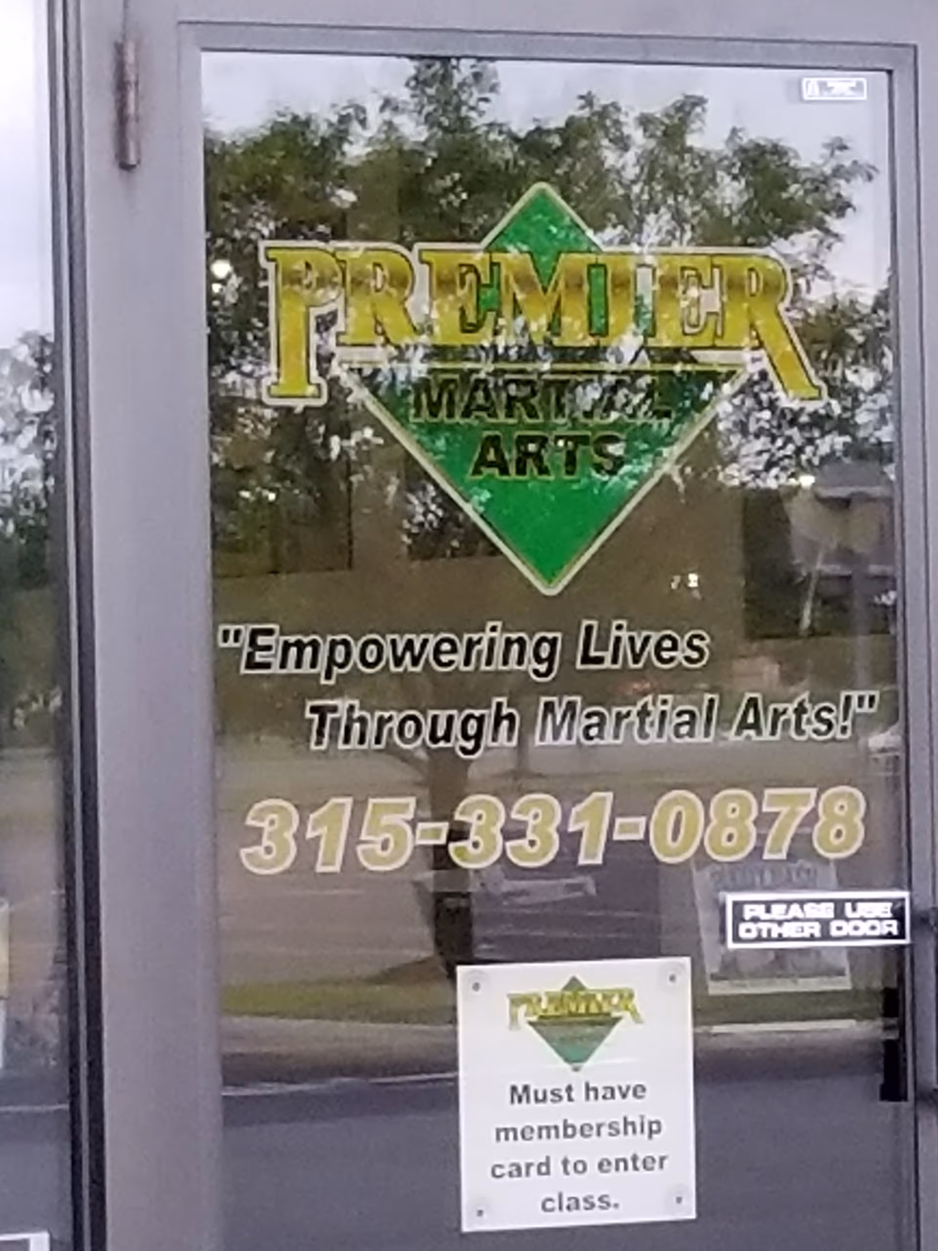 Glass door features Premier Martial Arts signage and a membership note, with trees reflected.