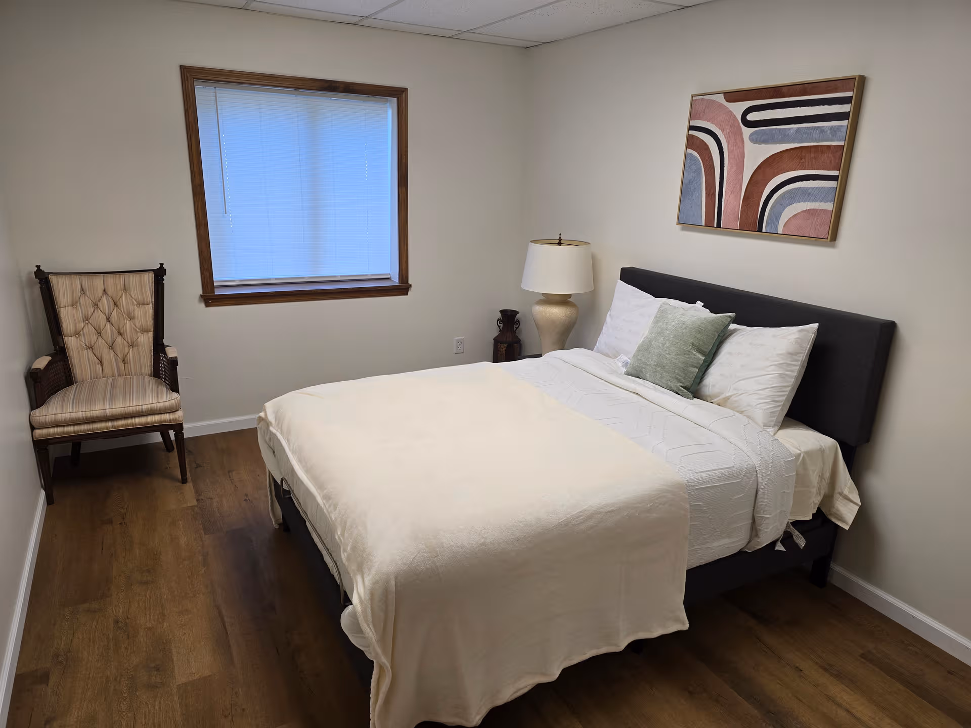 A neatly made bedroom with a bed, chair, window, and abstract wall art.