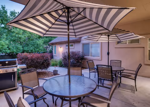 Outdoor patio with striped umbrellas, glass tables, chairs, and a barbecue grill.