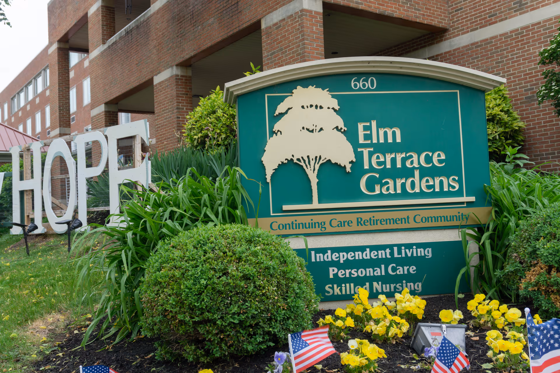 Elm Terrace Gardens exterior sign with greenery and yellow flowers in front of a brick building.
