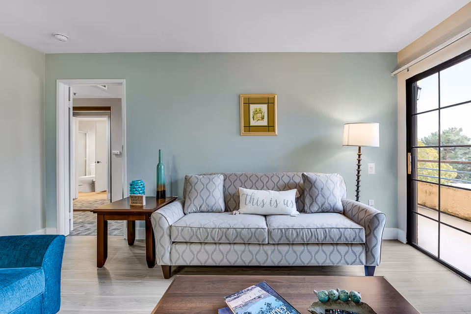 Bright living room with patterned sofa, side table, lamp, and balcony view.