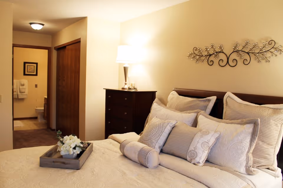 A cozy bedroom with a neatly made bed featuring multiple decorative pillows and a tray with white flowers. A wooden dresser with a lamp is beside the bed, and an open doorway leads to a bathroom with visible toilet and towels.