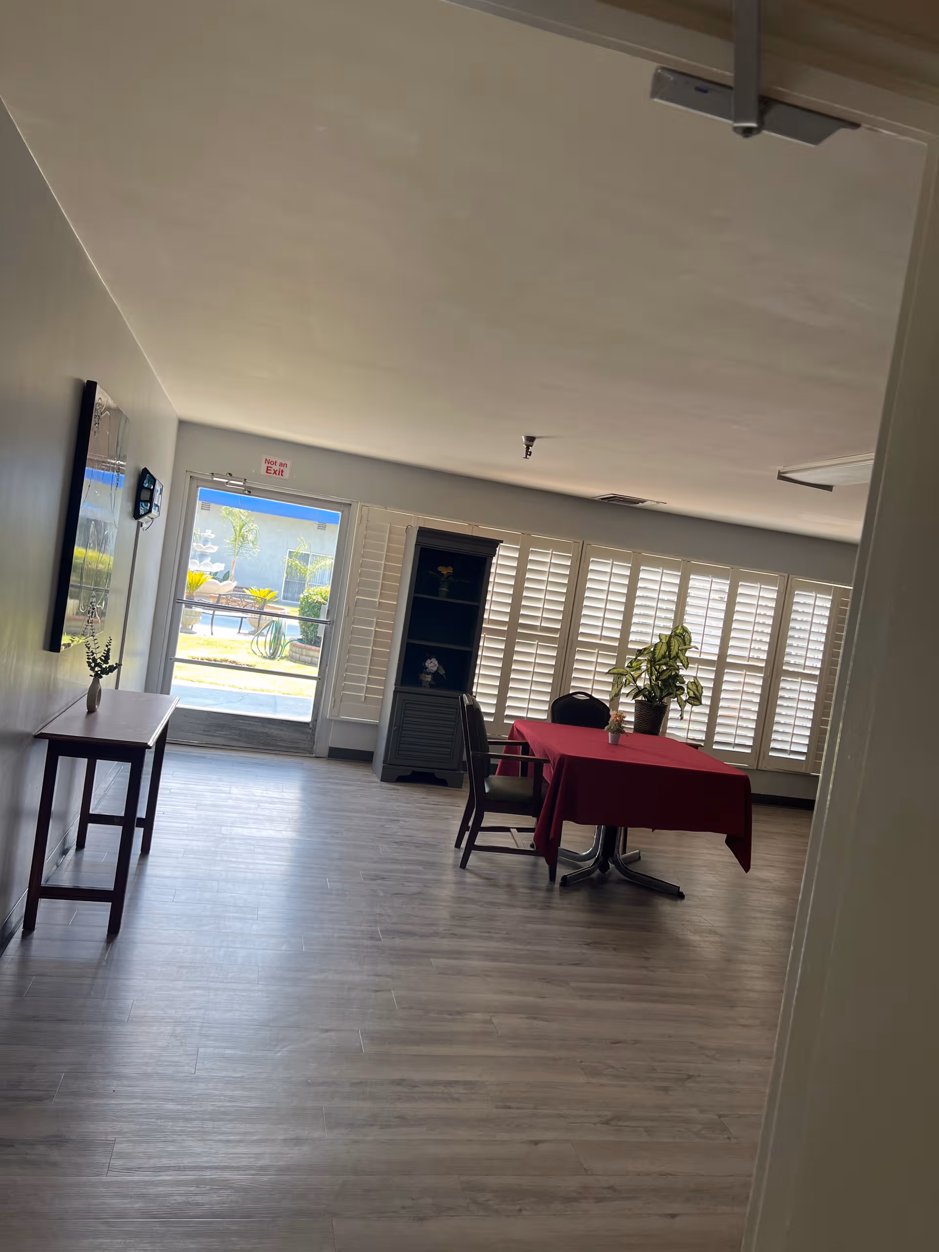 Empty dining area with a red tablecloth, chairs, plant, and large window shutters.