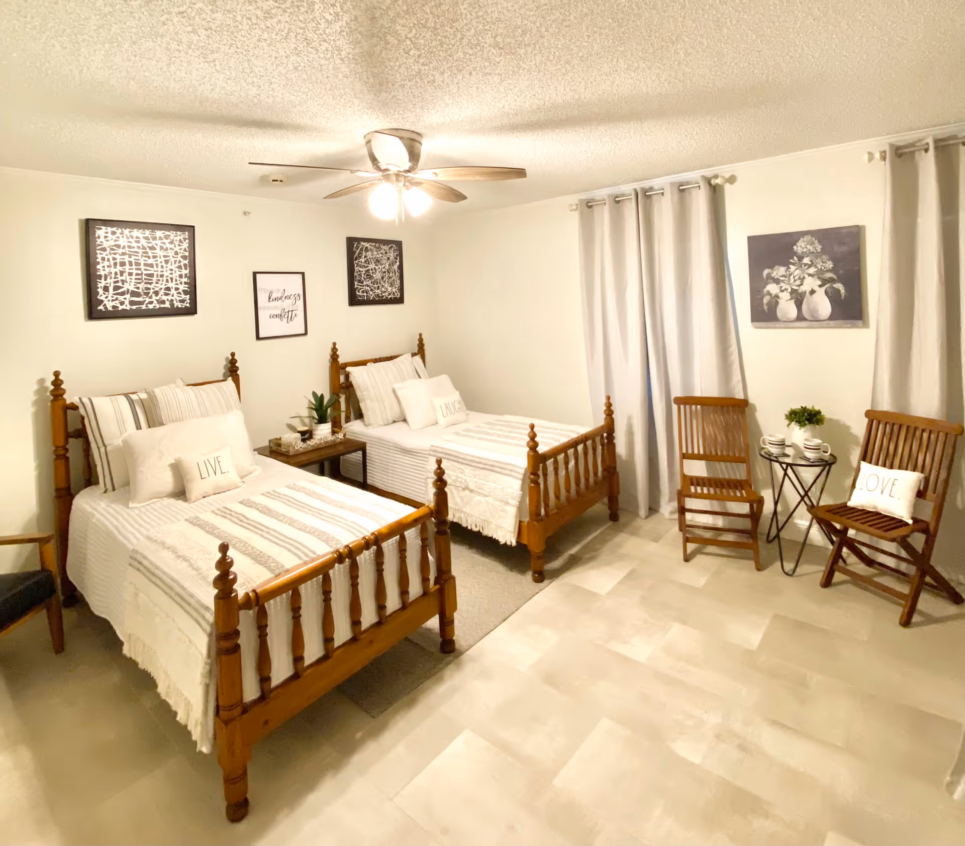 A bright bedroom with two wooden twin beds, each with white and beige striped bedding and decorative pillows labeled LIVE and LAUGH. Between the beds is a small wooden nightstand with a plant and decor. On the walls above the beds are three framed artworks, including one with the phrase 'kindness confetti.' To the right, two wooden chairs with a small round table between them hold two cups and a small plant. Light-colored curtains cover the windows, and a ceiling fan with lights is mounted on the ceiling.