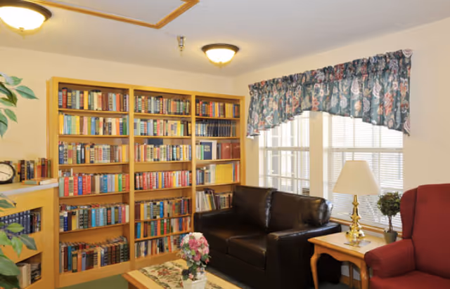 Cozy sitting room with wooden bookshelves, a dark leather sofa, an upholstered armchair, a coffee table with flowers, and a window with a floral valance.