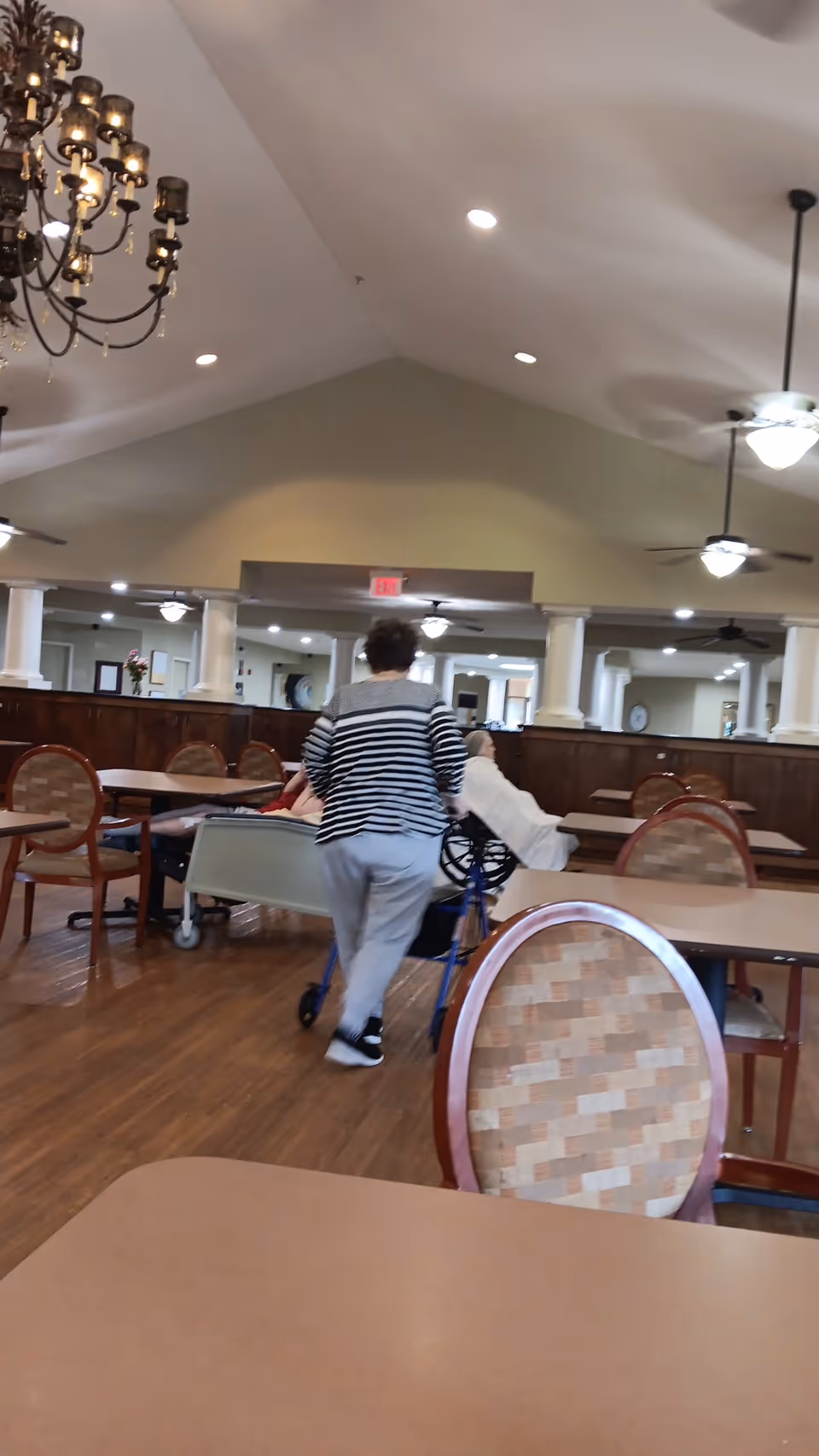 A person pushes a wheelchair through a spacious dining room in a senior living facility.
