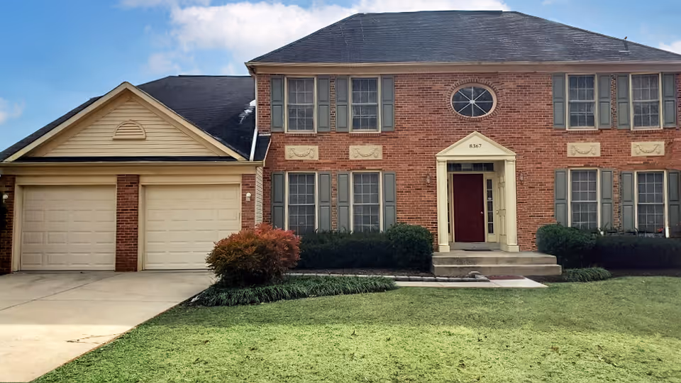 Front exterior view of a two-story brick house with multiple windows, a red front door with a small porch, two garage doors on the left side, and a well-maintained lawn with shrubs and a small tree in front.