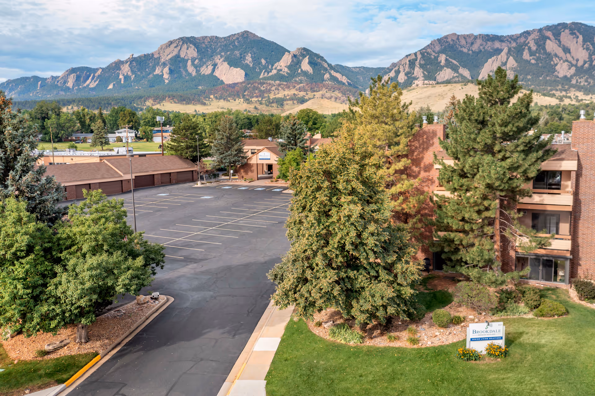 Exterior view of Brookdale Meridian Boulder senior living facility with a parking lot, trees, and mountains in the background under a partly cloudy sky.