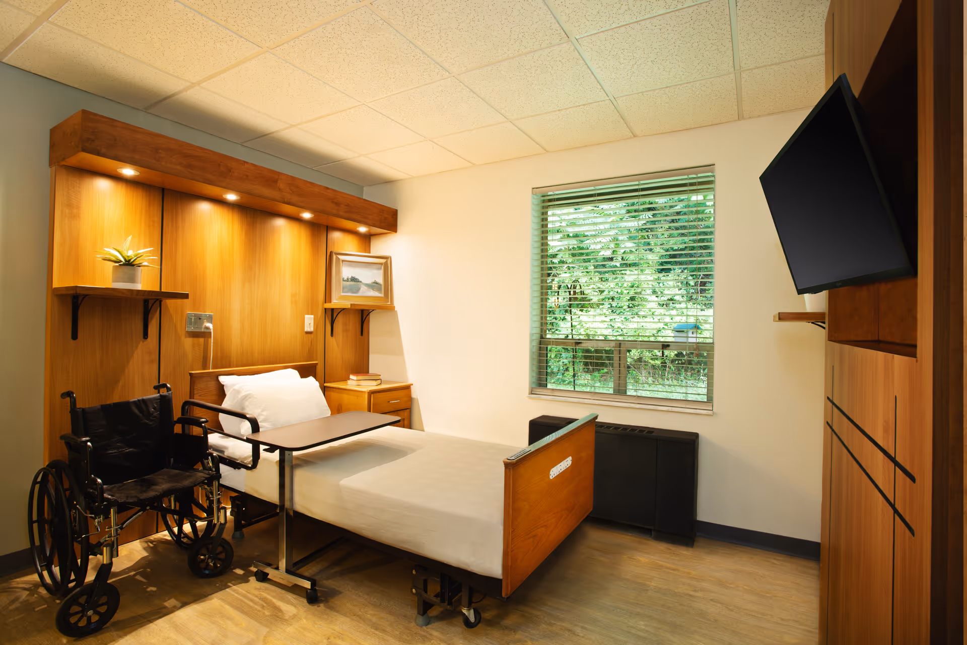 A nursing home bedroom with a single bed featuring a white pillow and sheets, a wooden headboard with built-in lighting, a small bedside table with books, a wheelchair positioned next to the bed, a window with blinds showing greenery outside, and a wall-mounted flat-screen TV.