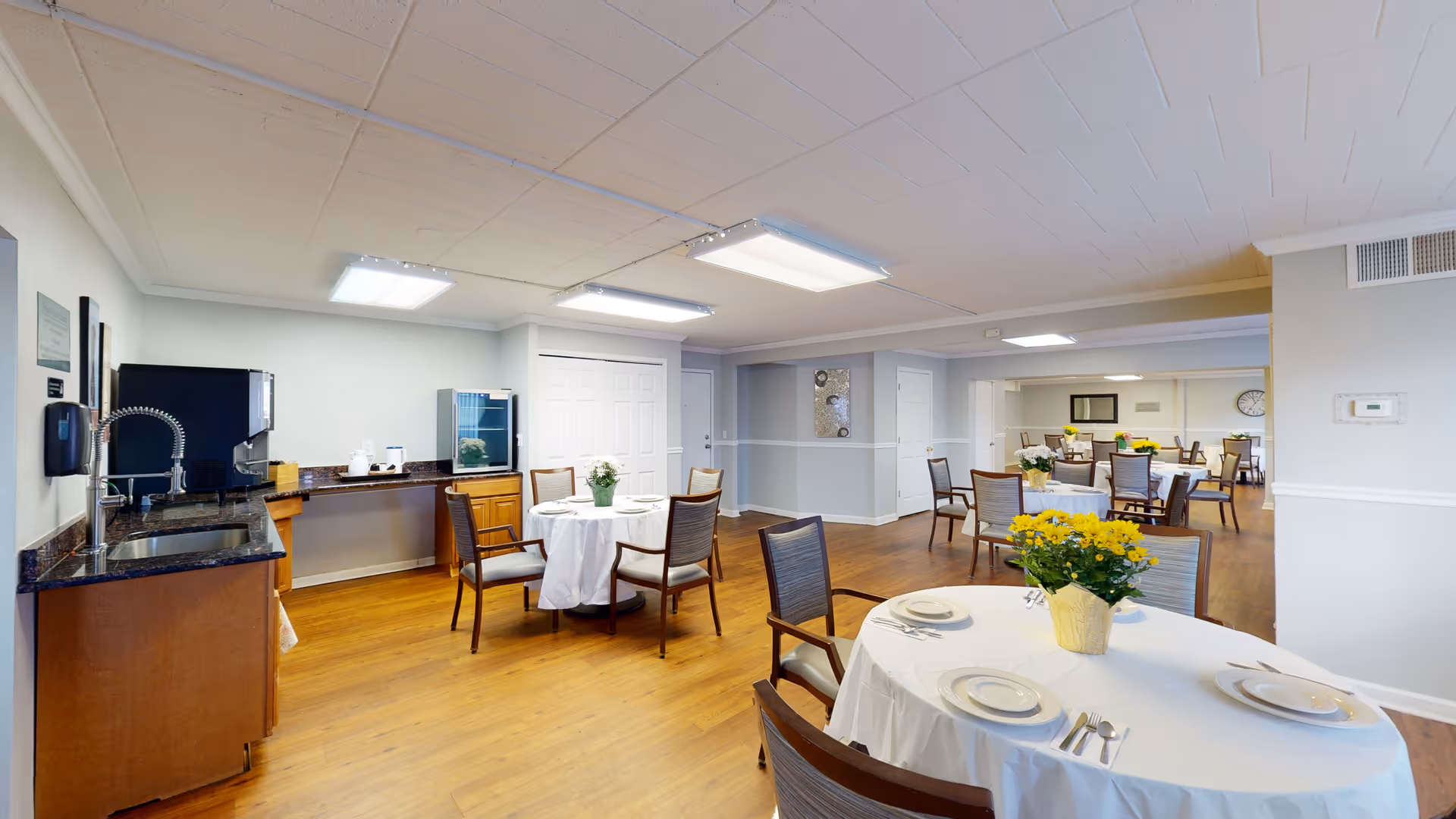 A spacious dining room in a senior living facility with multiple round tables covered with white tablecloths, each set with plates, utensils, and a flower centerpiece. The room has wooden flooring, light gray walls, and bright ceiling lights. There is a small kitchenette area with a sink, coffee machine, and a small refrigerator on the left side.
