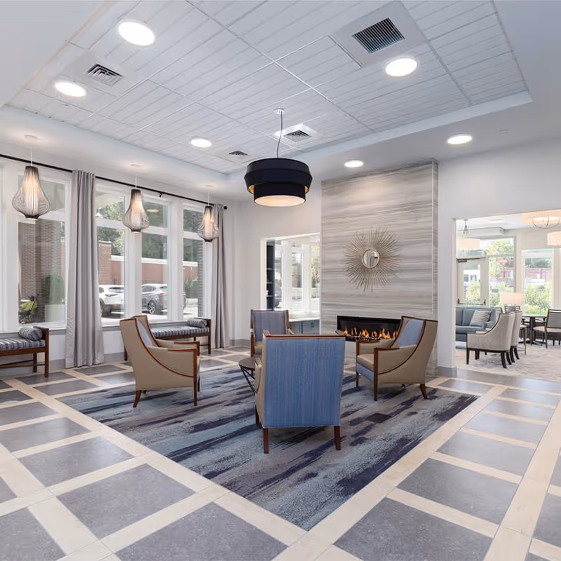 A modern senior living community lounge area with a central fireplace surrounded by four armchairs on a blue patterned rug. Large windows with gray curtains allow natural light to fill the space. Pendant lights hang from the ceiling, and additional seating is visible in an adjacent room.