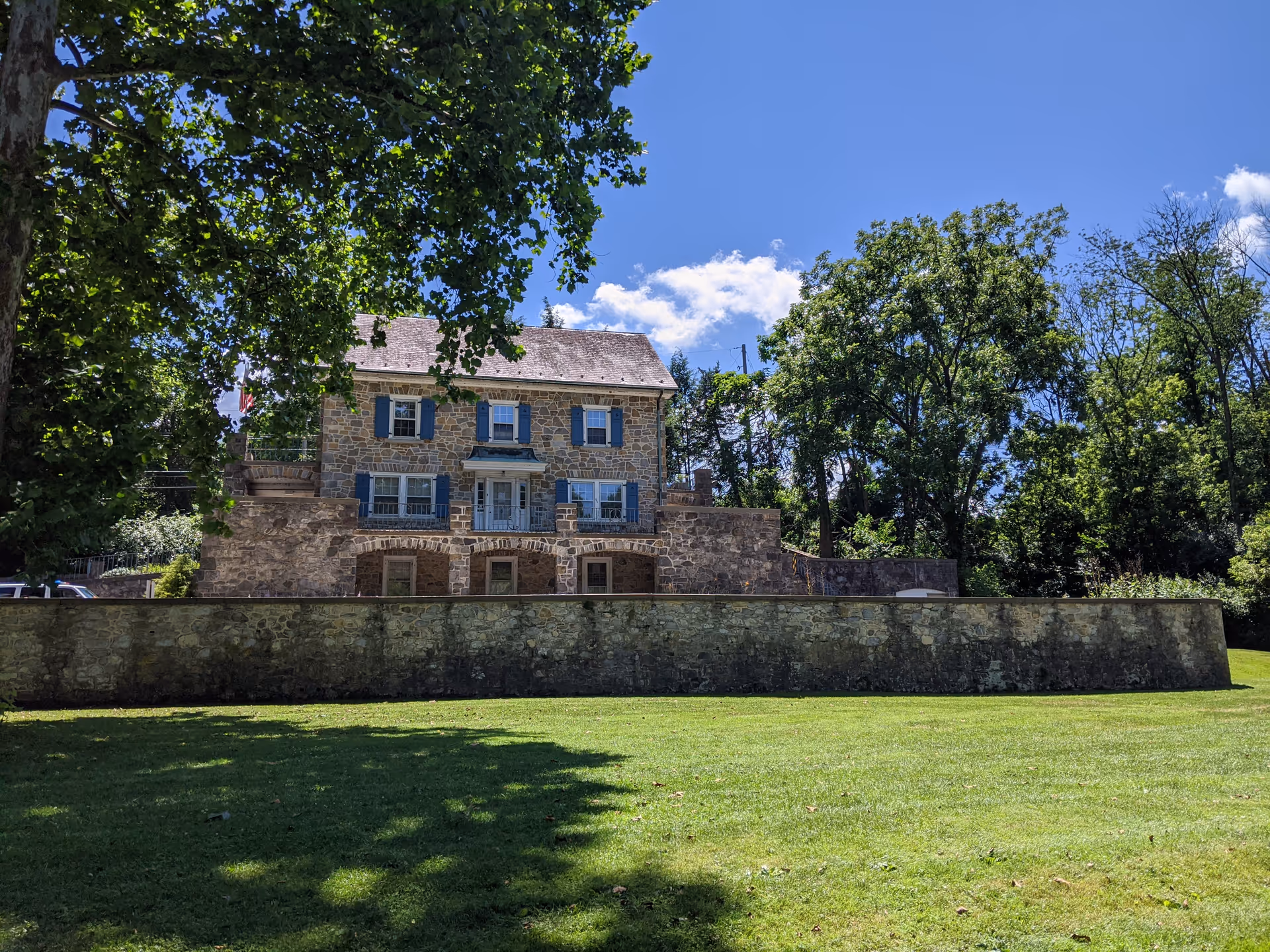 Stone two-story building with blue shutters sits behind a stone wall on a green lawn.