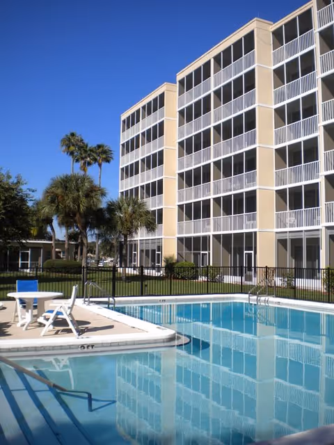 Outdoor swimming pool in front of a multi-story retirement building with screened balconies and palm trees under a clear blue sky.