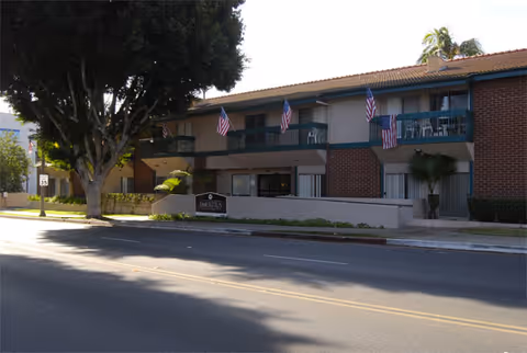 Exterior view of a two-story senior living facility building with balconies decorated with American flags. There is a large tree on the left side and a sign in front of the building that reads 'LINCOTUS'.