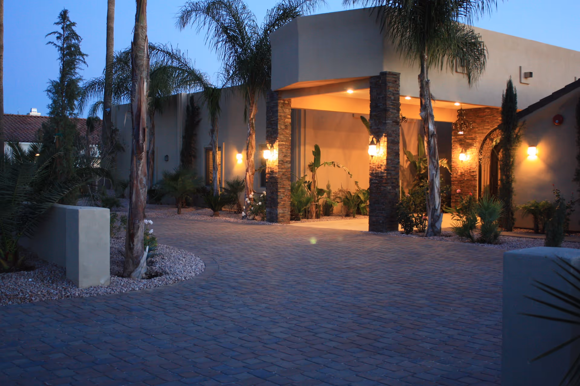 Evening view of the entrance to a building with stone pillars and warm lighting, surrounded by palm trees and desert landscaping with gravel and small plants.
