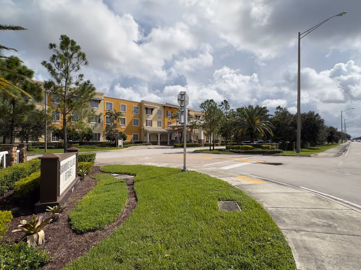 Front exterior view of a three-story yellow senior living building with landscaped grounds, driveway, and entrance sign.