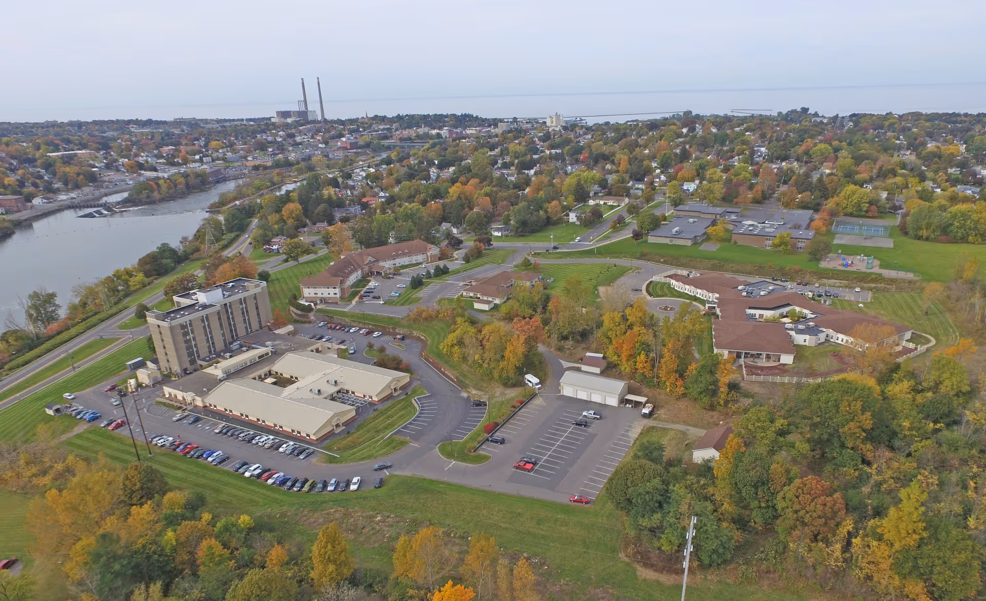 Aerial view of a senior living campus with several buildings, parking lots, roads, and trees showing fall colors near a river.