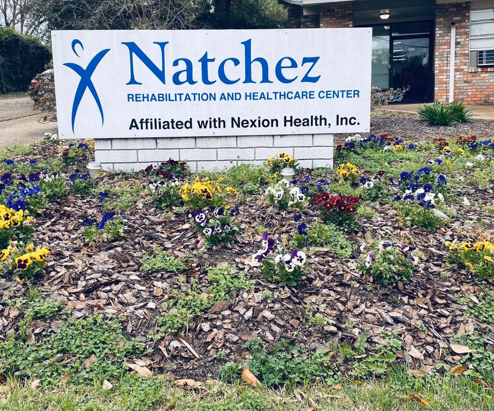 A white sign with blue and black text reading 'Natchez Rehabilitation and Healthcare Center Affiliated with Nexion Health, Inc.' is displayed in front of a building entrance. The sign is surrounded by a flower bed with colorful pansies and mulch. The building has a brick exterior and glass doors.
