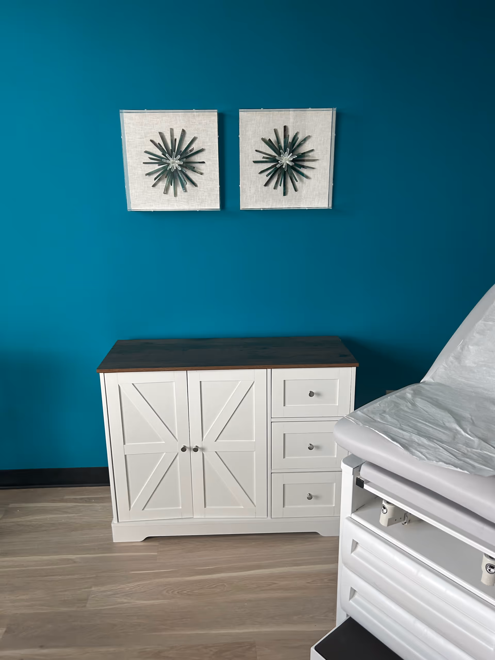 Teal-walled interior with white cabinet, framed art, and a medical examination table.