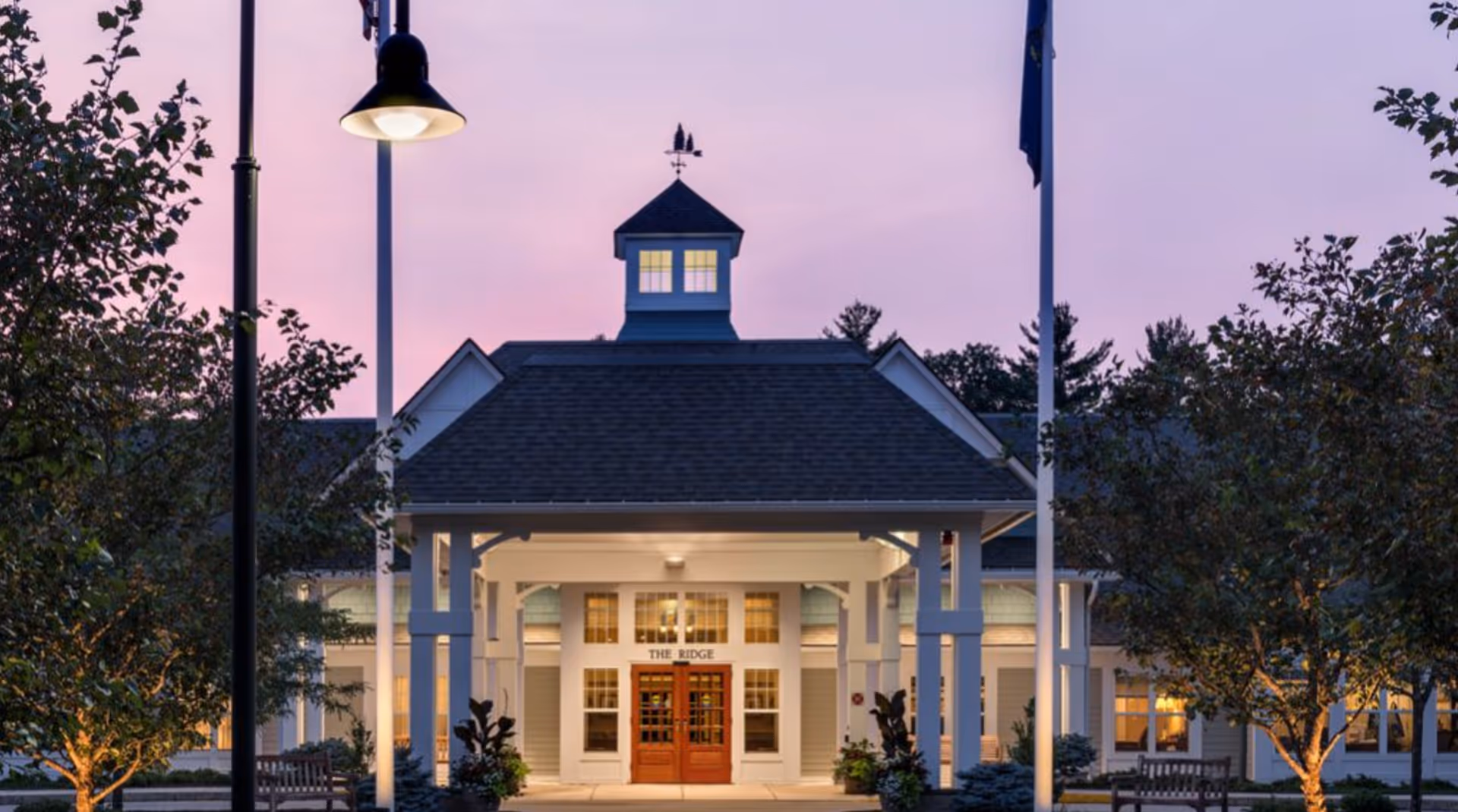 Front entrance of 'The Ridge' senior living building at dusk with an illuminated portico, cupola, flagpoles, and surrounding trees.