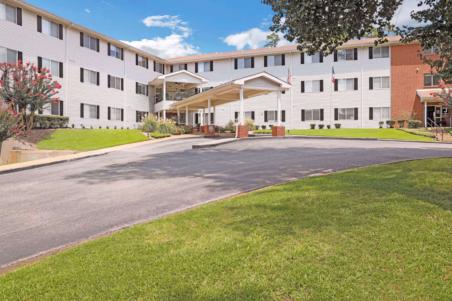 Exterior view of Holiday Colonial Village, a three-story senior living facility with white siding and brick accents. The building has multiple windows and a covered entrance with a driveway leading up to it. There is green grass and landscaping with trees and bushes around the building under a partly cloudy sky.