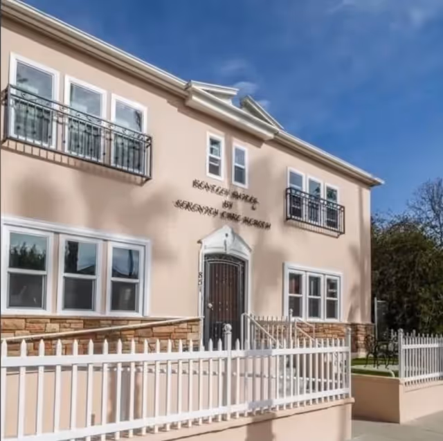 Two-story beige senior living building front with small balconies, a central entrance and a white picket fence under a blue sky.