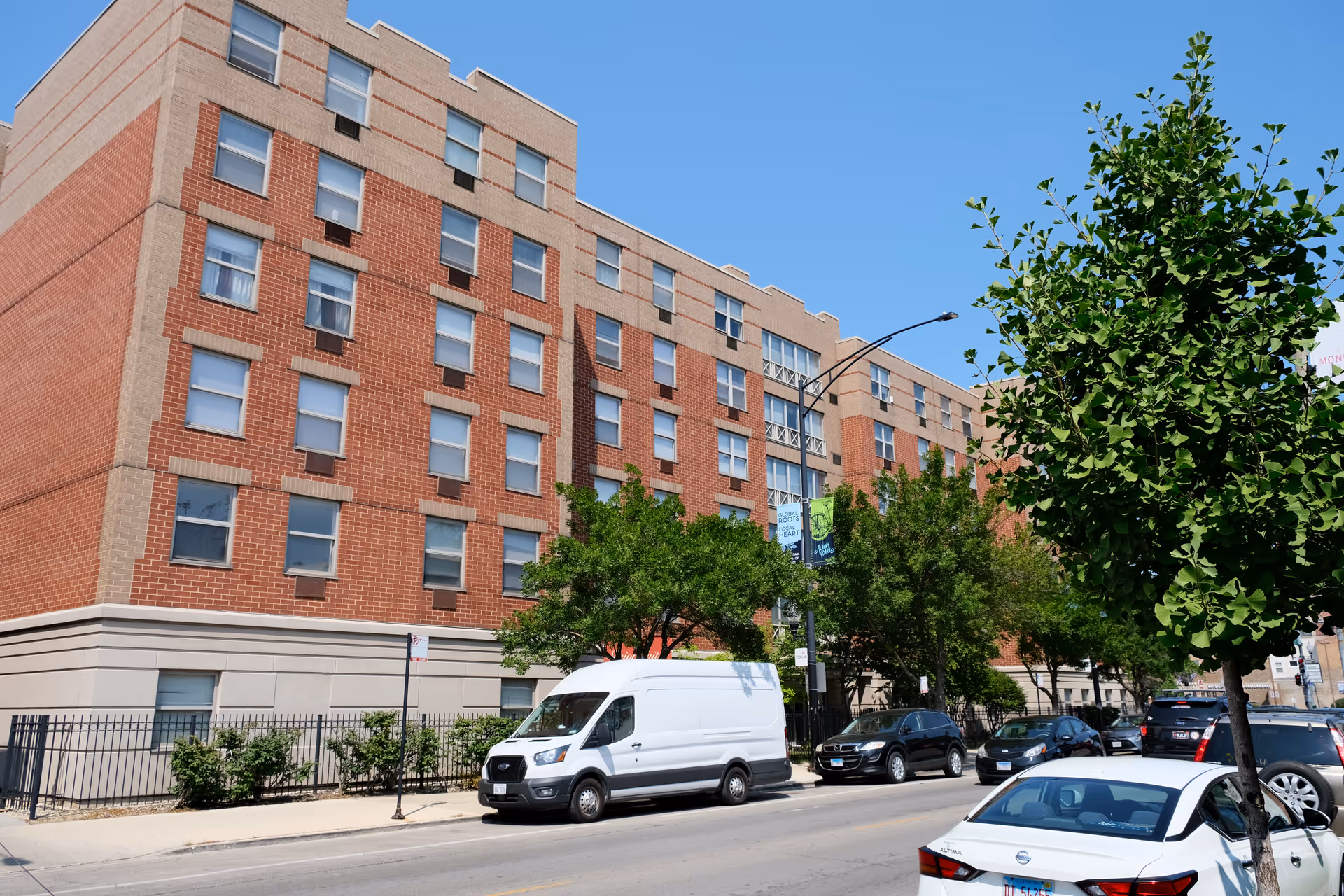 Brick multi-story residential building with trees and parked cars along a city street.
