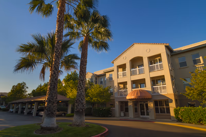 Exterior view of a three-story senior living facility building with balconies and an entrance covered by a brown awning. The scene includes tall palm trees, green bushes, and a covered parking area to the left under a clear blue sky.