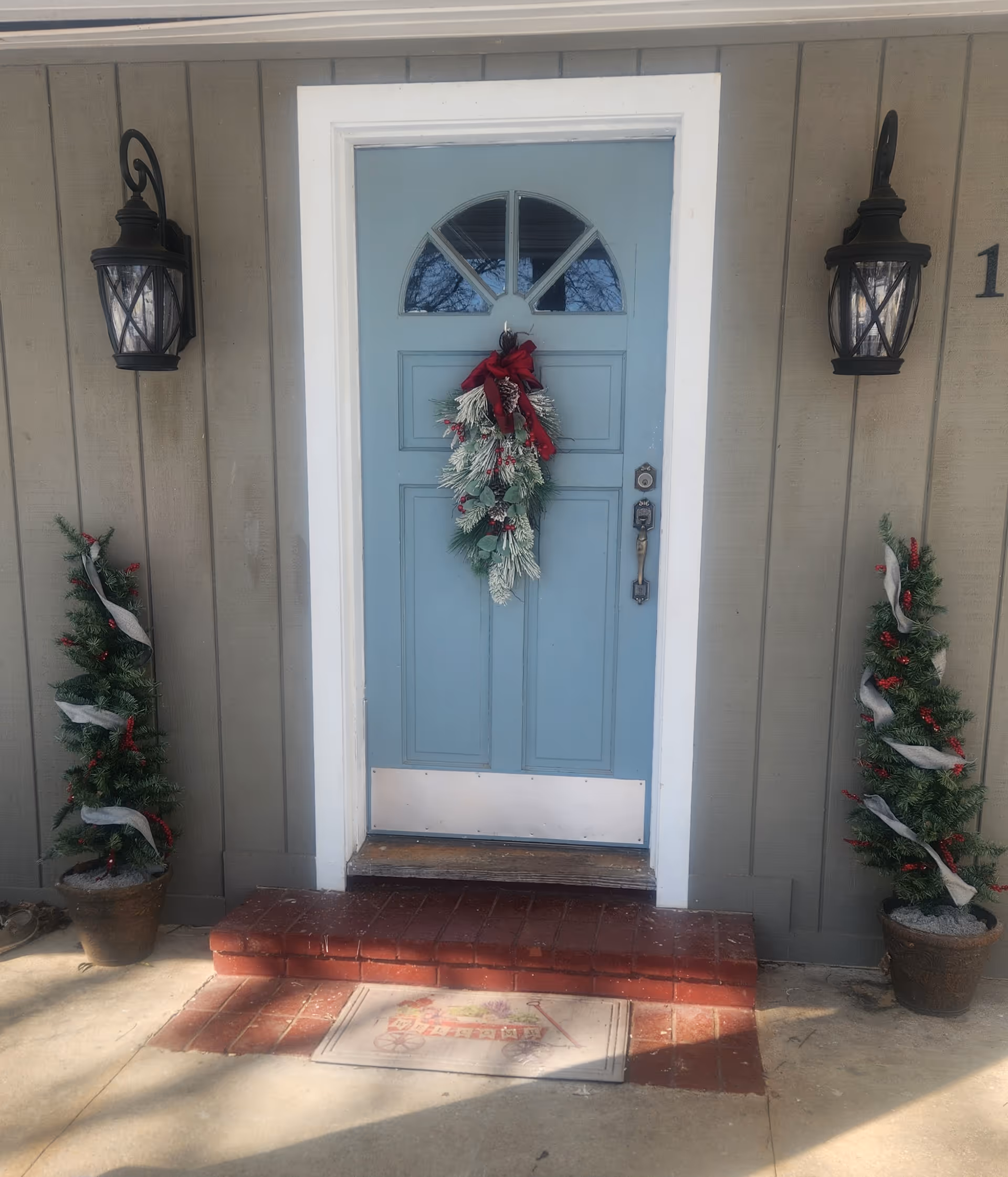 Blue front door with festive wreath, lanterns, and potted evergreens at entry.