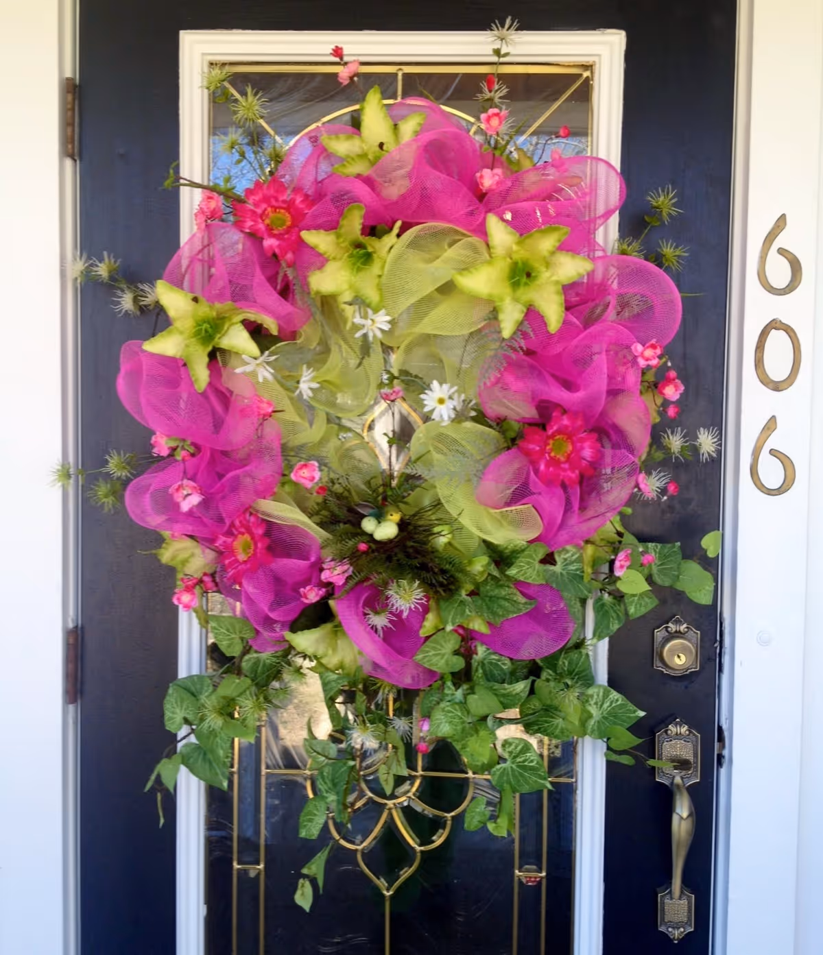 A black front door with a decorative glass panel and a large colorful wreath hanging on it. The wreath features bright pink and green mesh ribbons, green star-shaped flowers, small white daisies, pink flowers, and green leaves. The door has a brass handle and lock, and the house number 606 is displayed in gold on the white door frame.