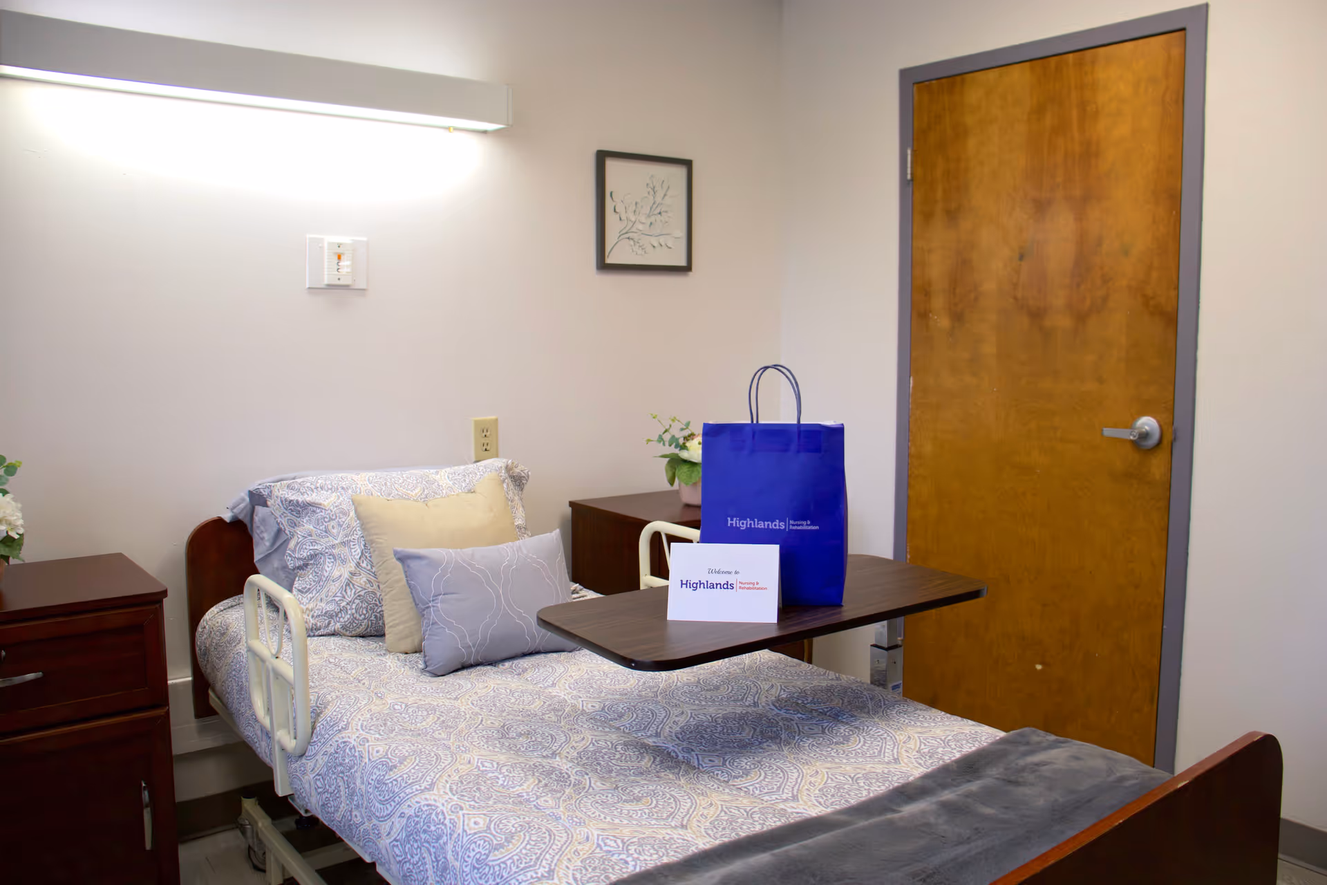 Hospital-style patient room with a bed, blue bedding, and a closed wooden door.