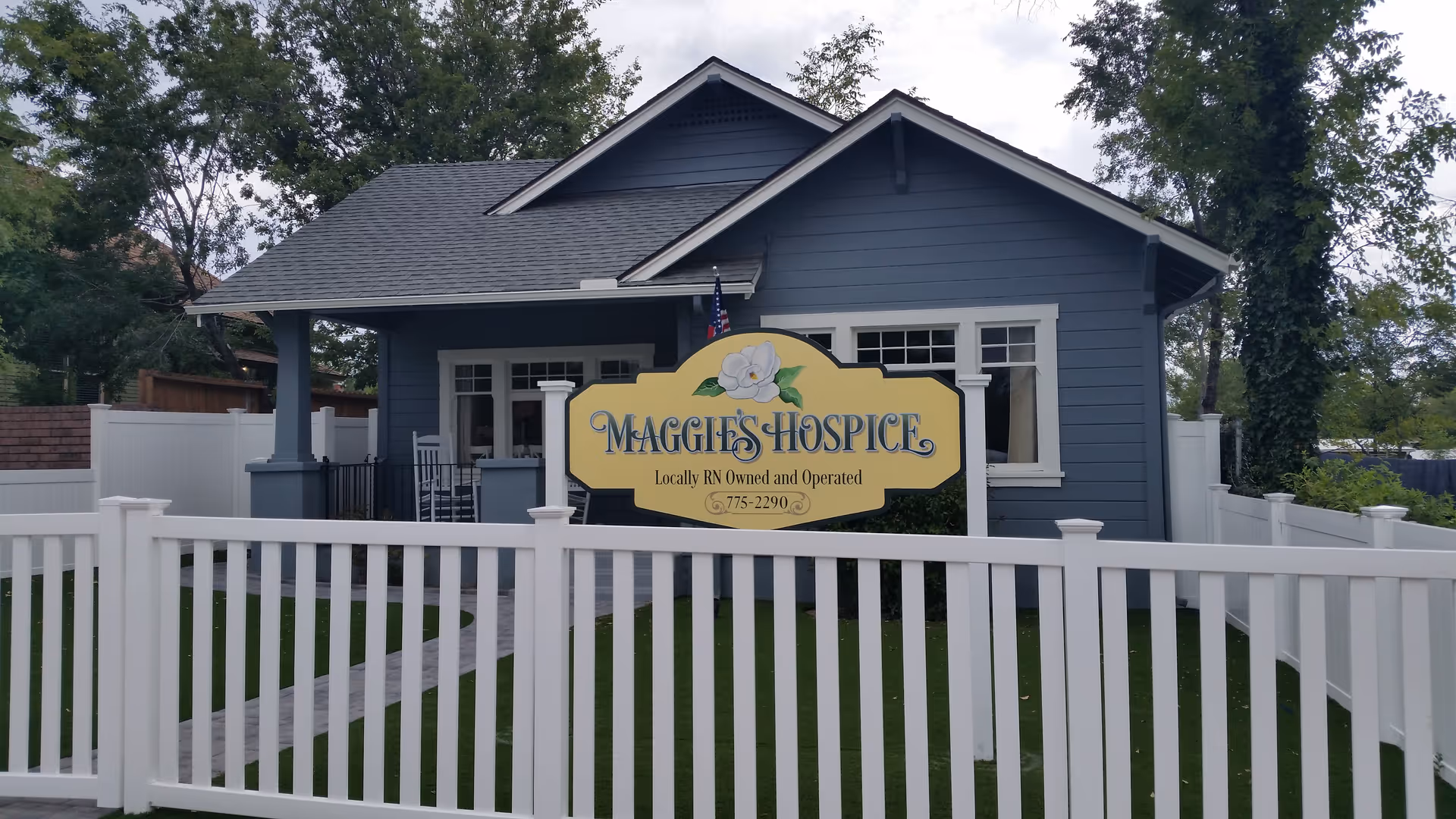 Blue single-story building behind a white fence with a Maggie's Hospice sign.